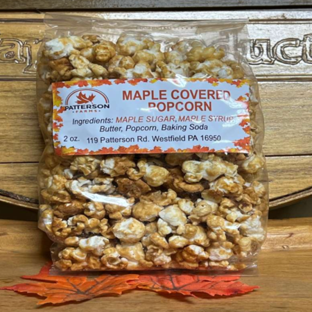 37366071-ceb9-4e7d-a86a-6647dcfd4d60 Bag of maple covered popcorn from Patterson Farms, featuring a clear label with ingredients including maple sugar, maple syrup, butter, popcorn, and baking soda. The package contains 2 ounces and is set against a wooden background with autumn leaves.