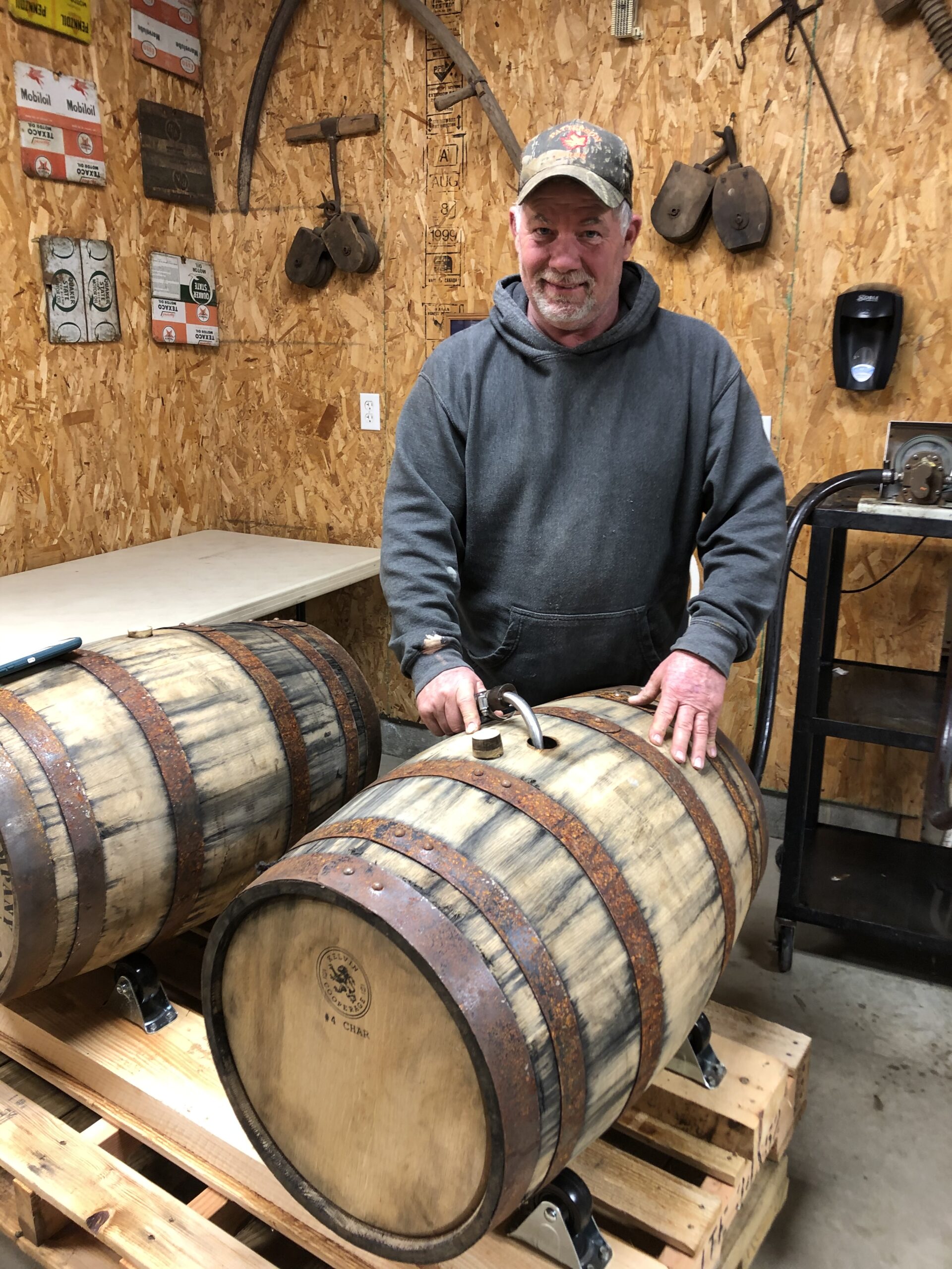 Man working with rustic wooden barrels in a workshop, surrounded by vintage signs and tools, showcasing craftsmanship in barrel making.