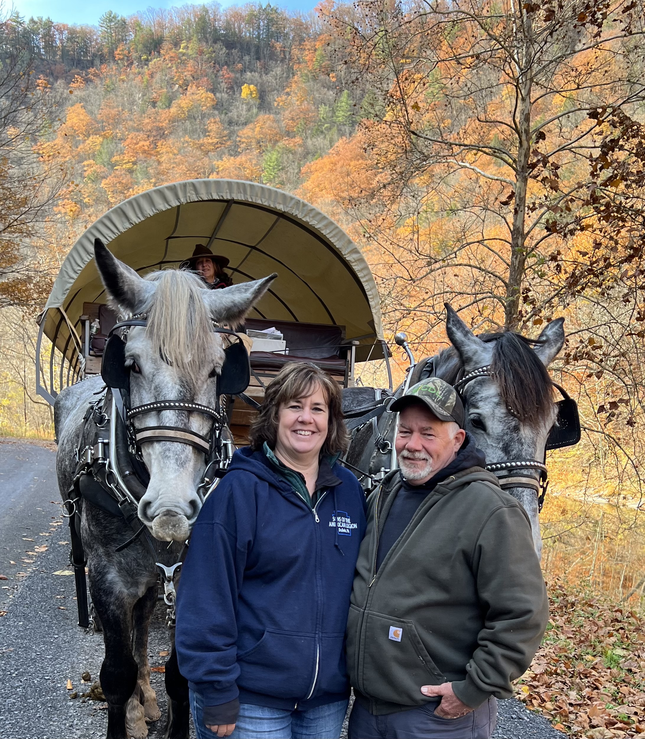 A couple stands in front of two horses hitched to a covered wagon, surrounded by vibrant autumn foliage in the background. The scene captures a picturesque outdoor moment, highlighting equestrian culture and the beauty of fall scenery.