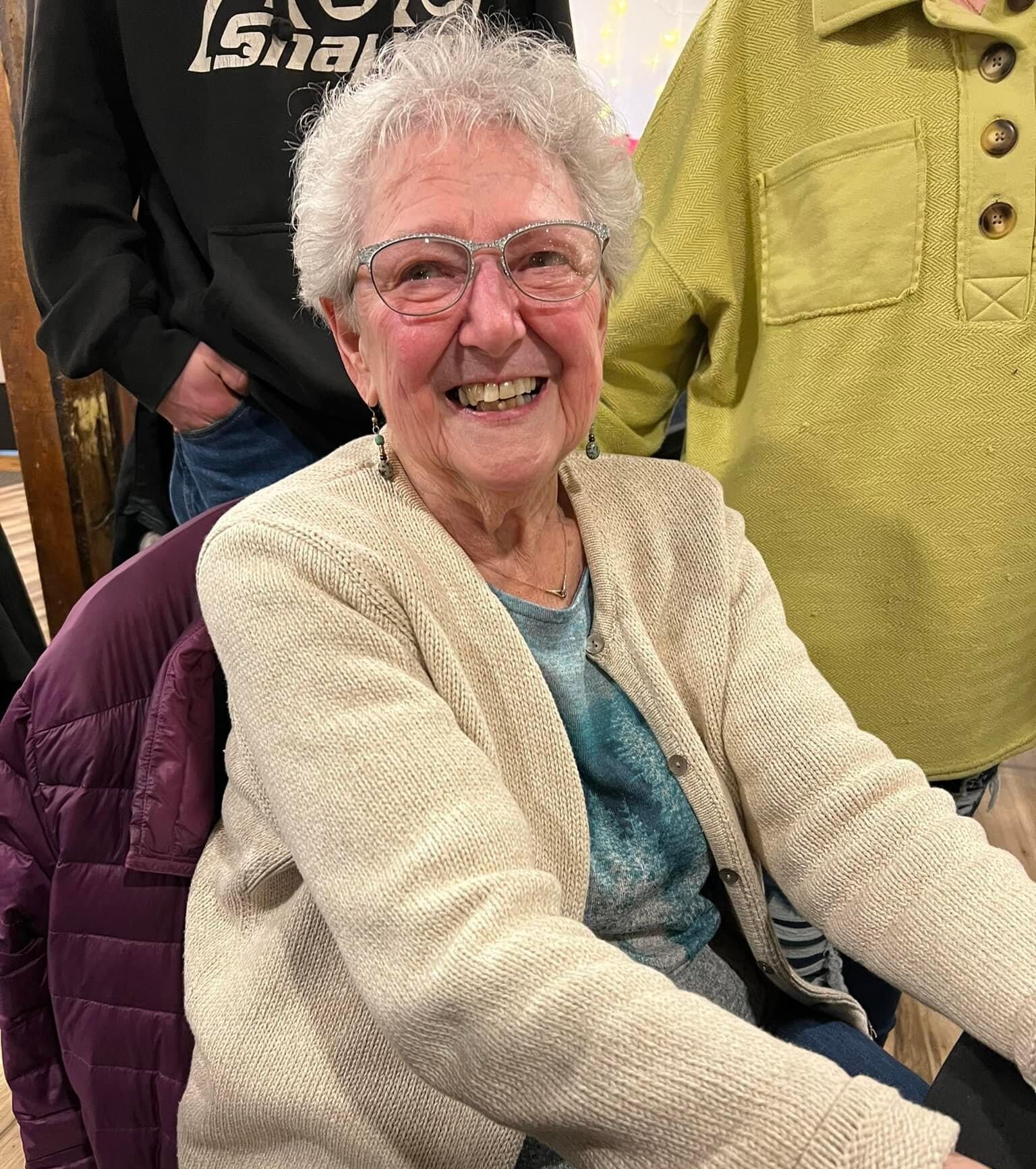 Smiling elderly woman with short, curly gray hair and glasses, wearing a beige cardigan and seated at a table. Surrounded by family members, she exudes warmth and joy in a cozy indoor setting.