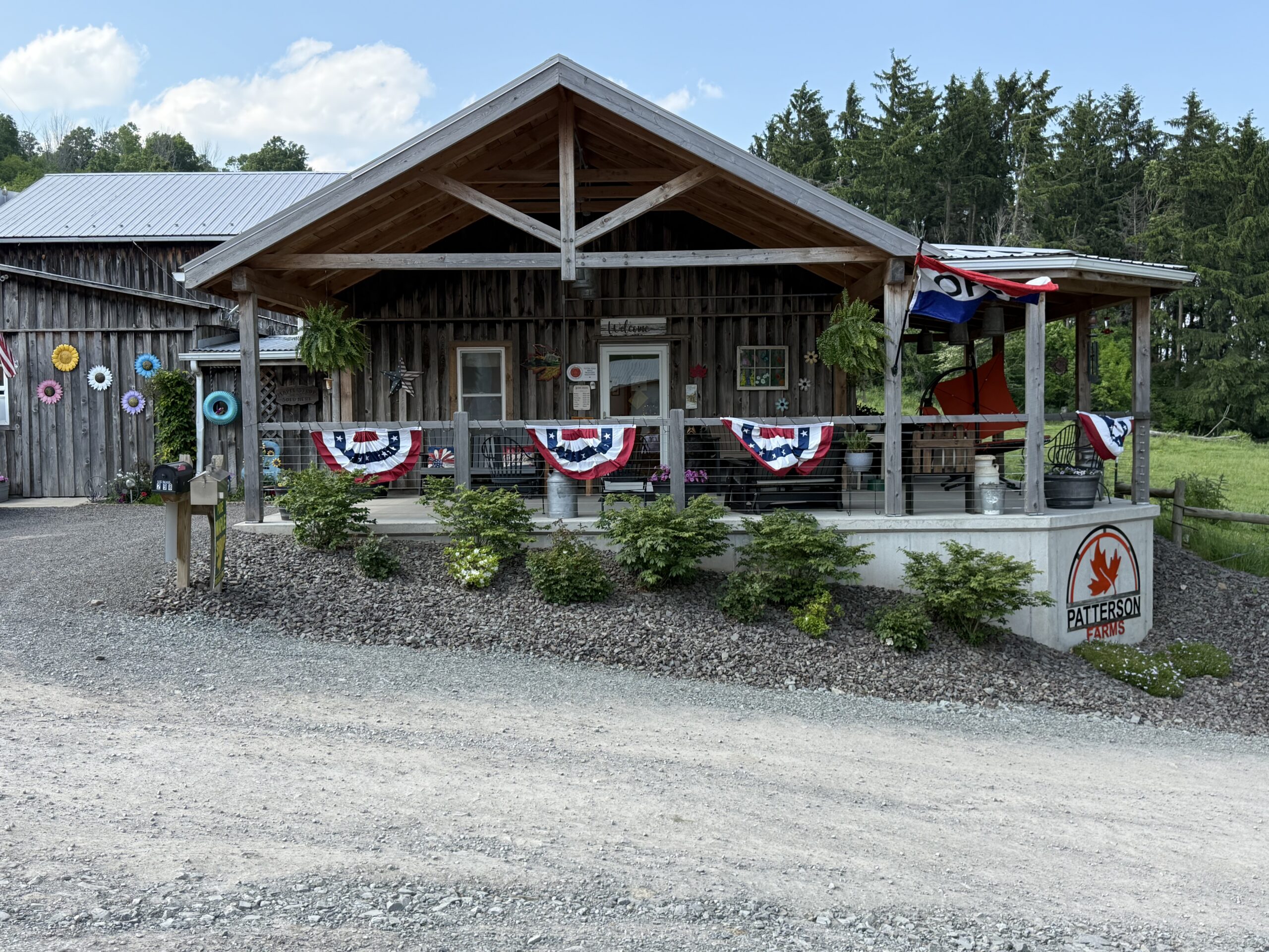 Exterior view of Patterson Farms, featuring a rustic wooden building with a welcoming porch adorned with red, white, and blue decorations, surrounded by greenery and colorful flower decorations.