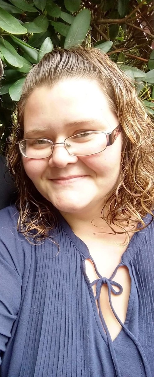 Smiling woman with curly hair wearing glasses and a navy blouse, posing outdoors with greenery in the background.