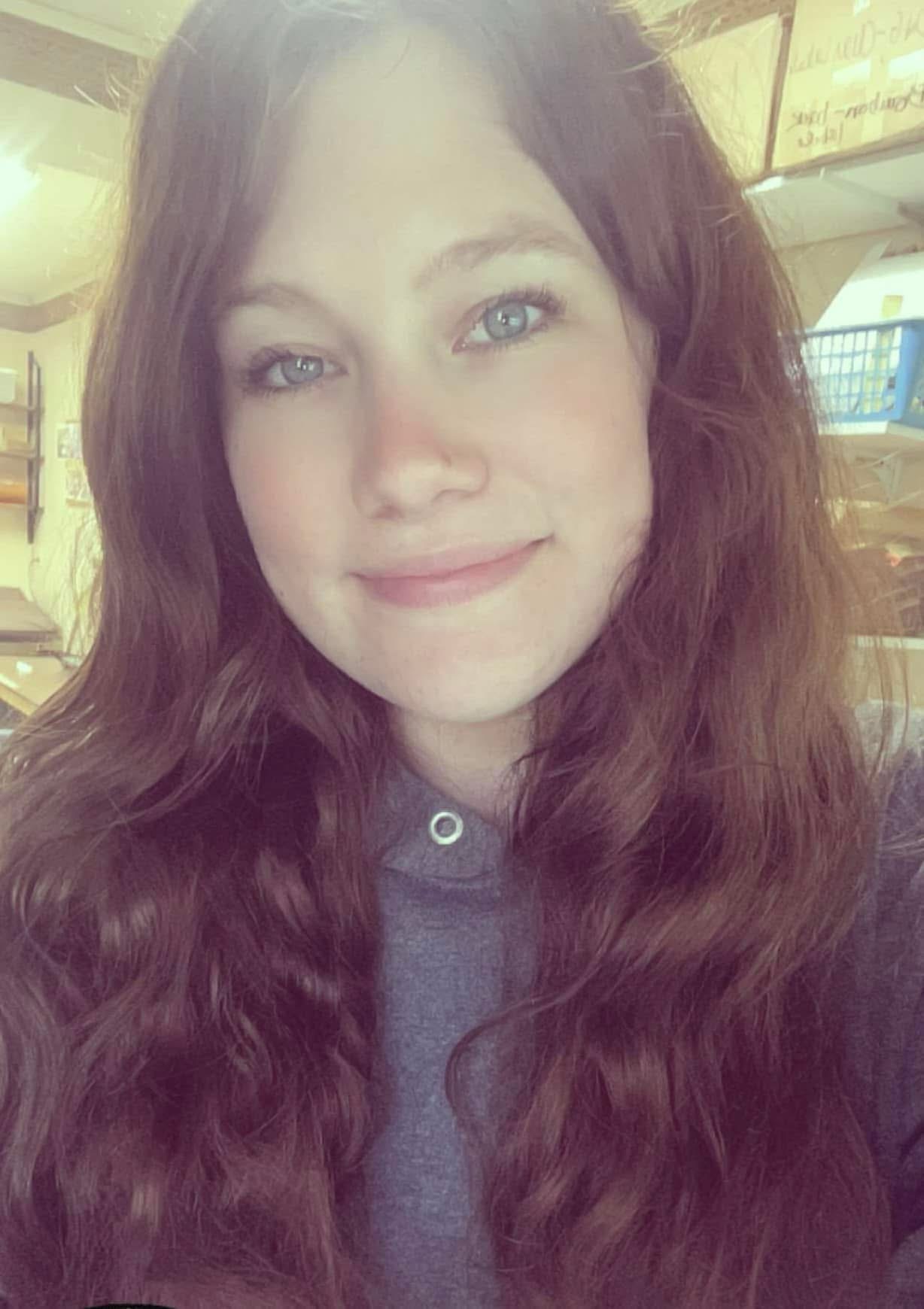 Young woman with long, wavy brown hair and green eyes smiling at the camera, wearing a gray hoodie. Soft lighting highlights her features, with a blurred background of shelves and storage.