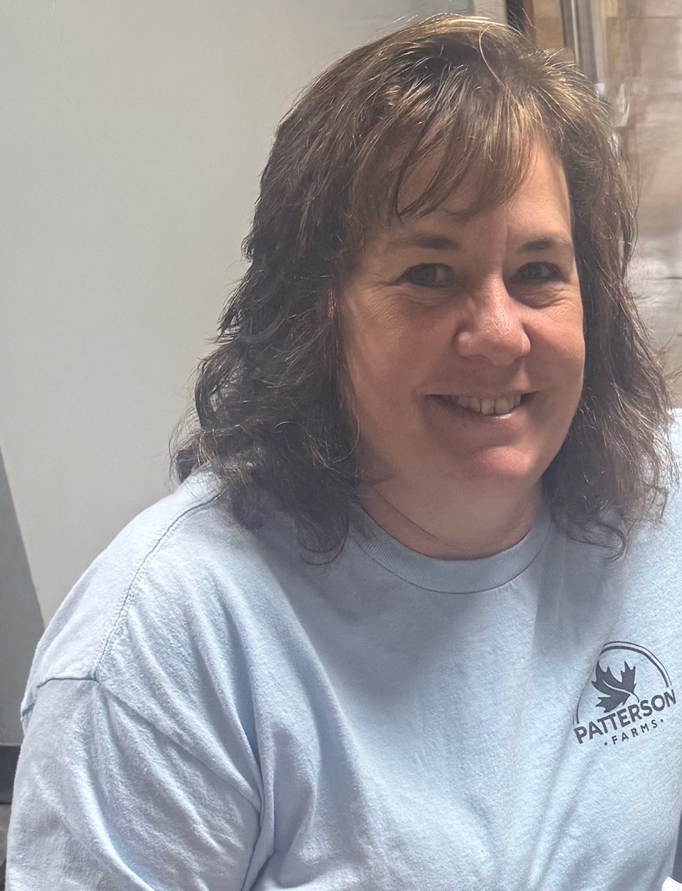 Smiling woman with shoulder-length brown hair wearing a light blue t-shirt with "Patterson Farms" logo, seated indoors.