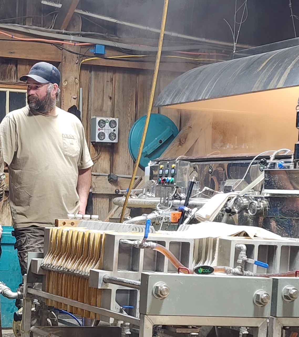 A worker in a workshop stands beside a large piece of equipment used for syrup production, with steam rising in the background. The setting features wooden walls and various control panels, indicating an active manufacturing environment.