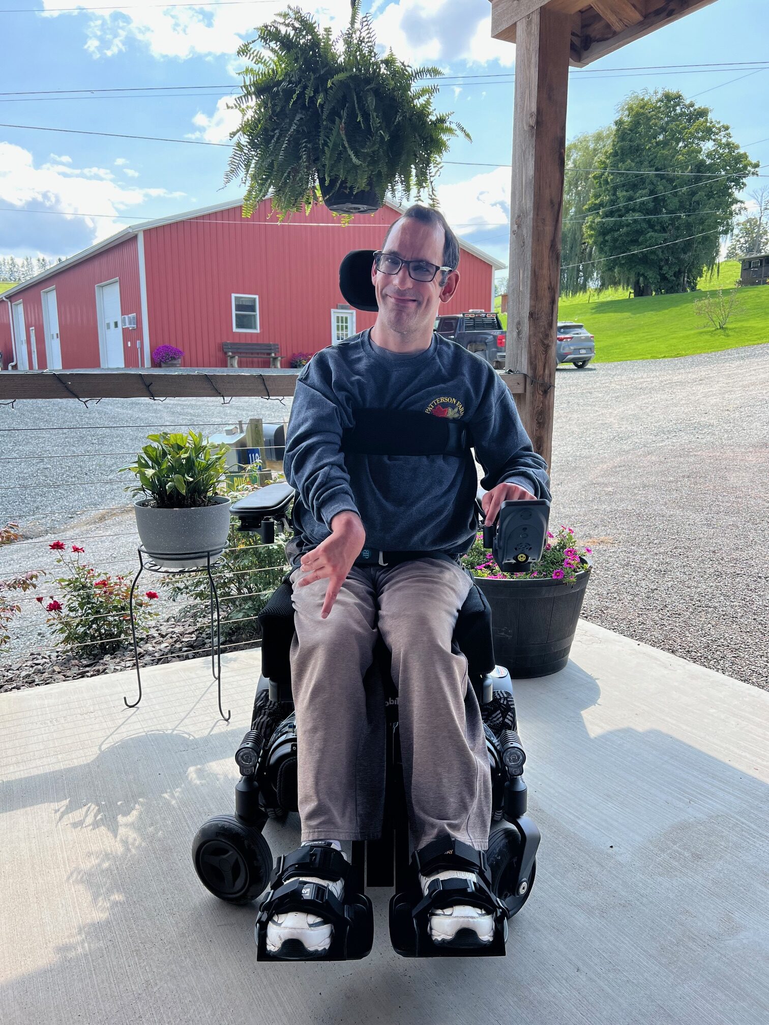 Man in a power wheelchair, wearing glasses and a dark sweatshirt, smiles while sitting on a porch with potted plants and a red barn in the background. The scene captures a sunny day with greenery and flowers.