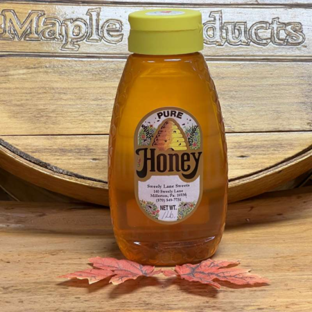 aca8a217-654b-4409-913a-88b2932c4862 Bottle of pure honey from Sweet Lane Sweets, 1 lb, displayed on a wooden surface with a maple products sign in the background and autumn leaves in the foreground.