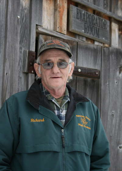 Elderly man wearing glasses and a green jacket stands in front of a wooden door marked "Evaporator Room," showcasing a rustic setting related to maple syrup production.